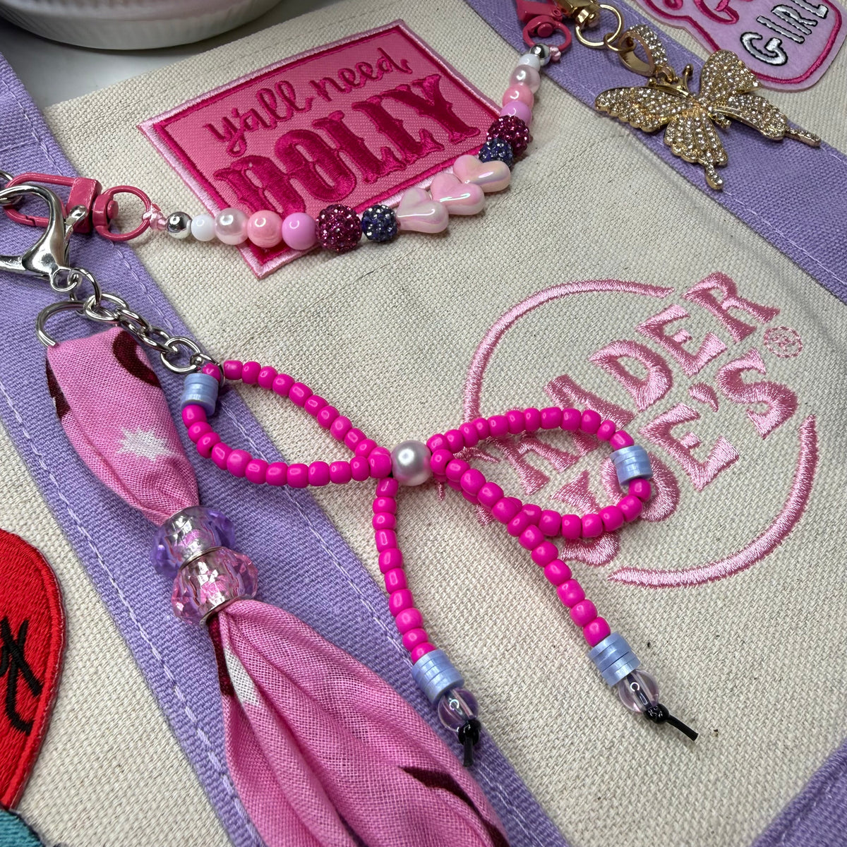 A pink beaded keychain with a bow charm, silver clasp, and tassel, displayed on a beige canvas bag with embroidered patches. Dolly Parton You Go Girl mini tote by PrettyMUCH.