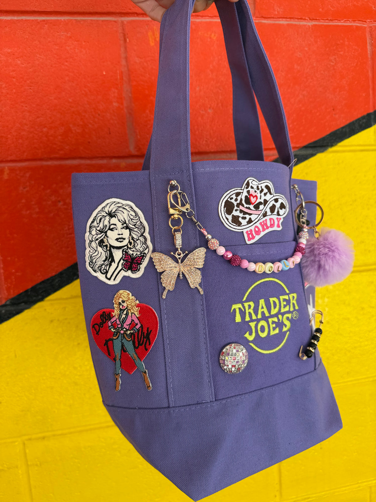 A purple Trader Joe's mini tote decorated with patches, pins and charms, including the embroidered Trader Joe's logo, a pink SWEET BUT STRONG banner patch and a rhinestone butterfly charm, held against a bright yellow and orange painted brick wall. Dolly Parton Sweet but Strong mini tote by PrettyMUCH.