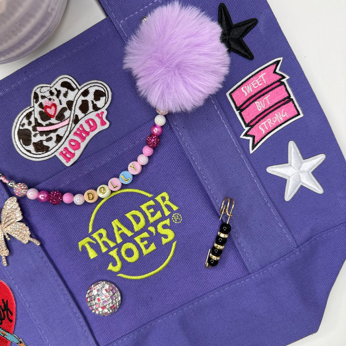 A close-up of a purple fabric tote bag decorated with multiple patches, a lavender pom-pom, a beaded bracelet, a black starfish patch, a white star patch, and a yellow Trader Joe's logo. Dolly Parton Sweet but Strong mini tote by PrettyMUCH.