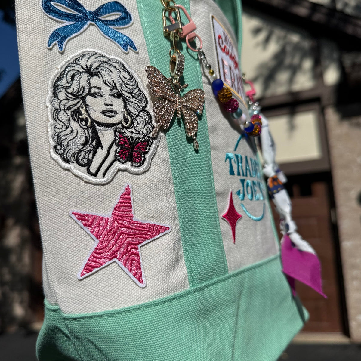 A close-up of a personalized canvas tote bag with mint green and off-white panels, decorated with embroidered patches, a butterfly charm, and colorful beads, photographed outdoors in front of a building. Dolly Parton Cowgirl Era mini tote by PrettyMUCH.