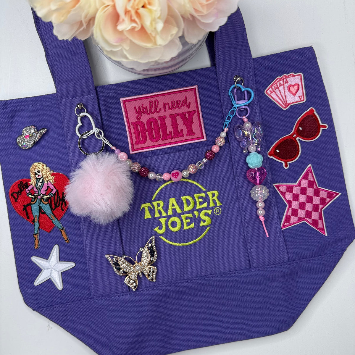A purple Trader Joe's mini tote decorated with patches, keychains and charms, including a pink pom-pom, the Trader Joe's logo and the hot pink 'y'all need DOLLY' patch, displayed on a white surface with a flower in the background. Dolly Parton Aces High mini tote by PrettyMUCH.