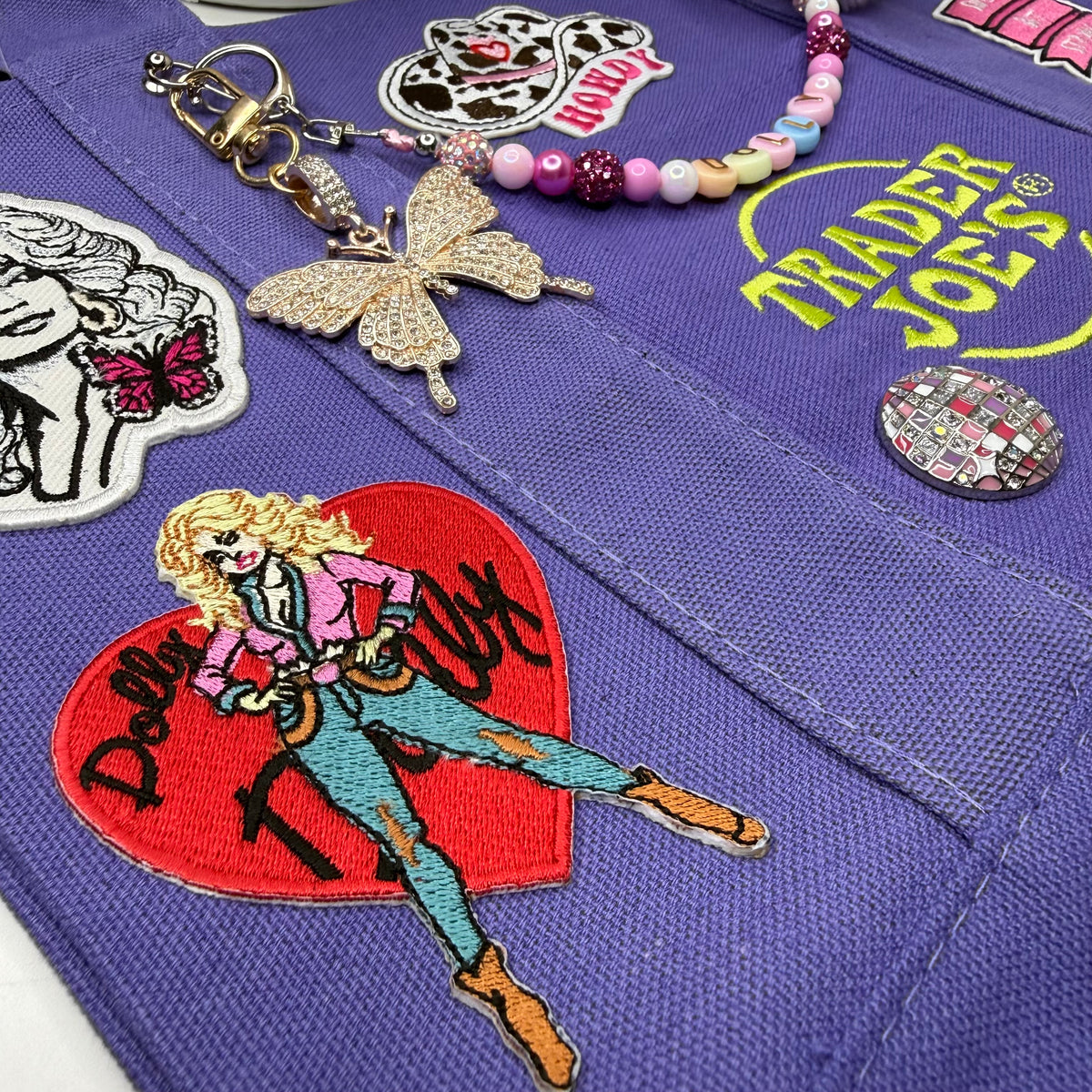 Close-up of the purple Trader Joe's mini tote with the embroidered Dolly Parton red heart patch, a gold rhinestone butterfly charm, a beaded DOLLY bag chain and the yellow embroidered Trader Joe's logo. Dolly Parton Sweet but Strong mini tote by PrettyMUCH.