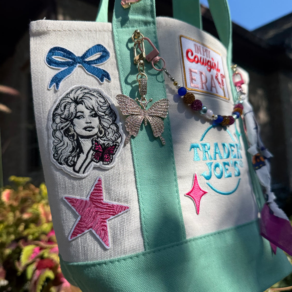 A mint green and white canvas tote bag decorated with colorful embroidered patches, a butterfly charm, and a beaded keychain, photographed outdoors with blurred foliage in the background. Dolly Parton Cowgirl Era mini tote by PrettyMUCH.