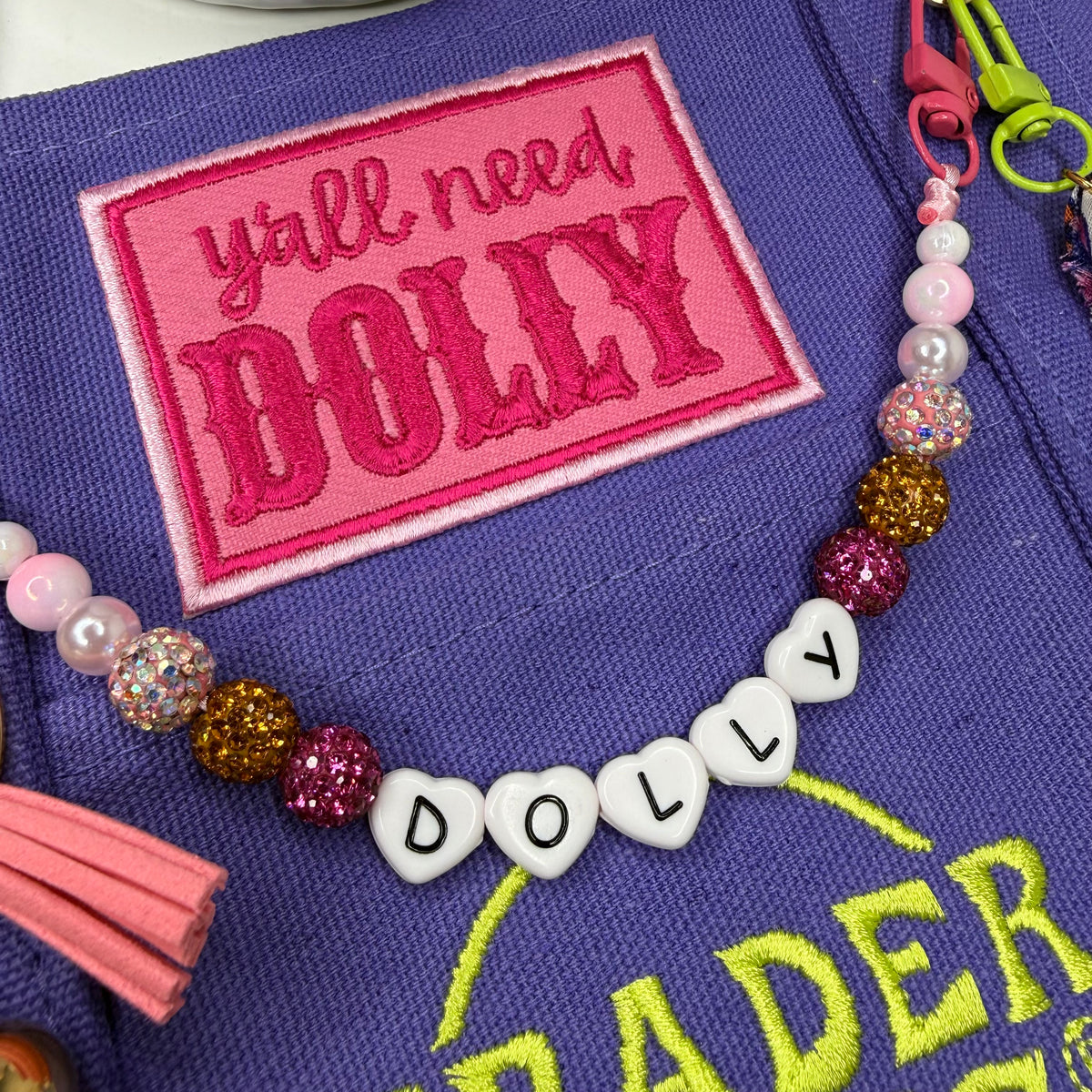 Close-up of the purple Trader Joe's mini tote showing the hot pink 'y'all need DOLLY' patch and the beaded DOLLY bag chain in white heart letter beads, above the green embroidered Trader Joe's logo. Dolly Parton Y'all Need Dolly mini tote by PrettyMUCH.