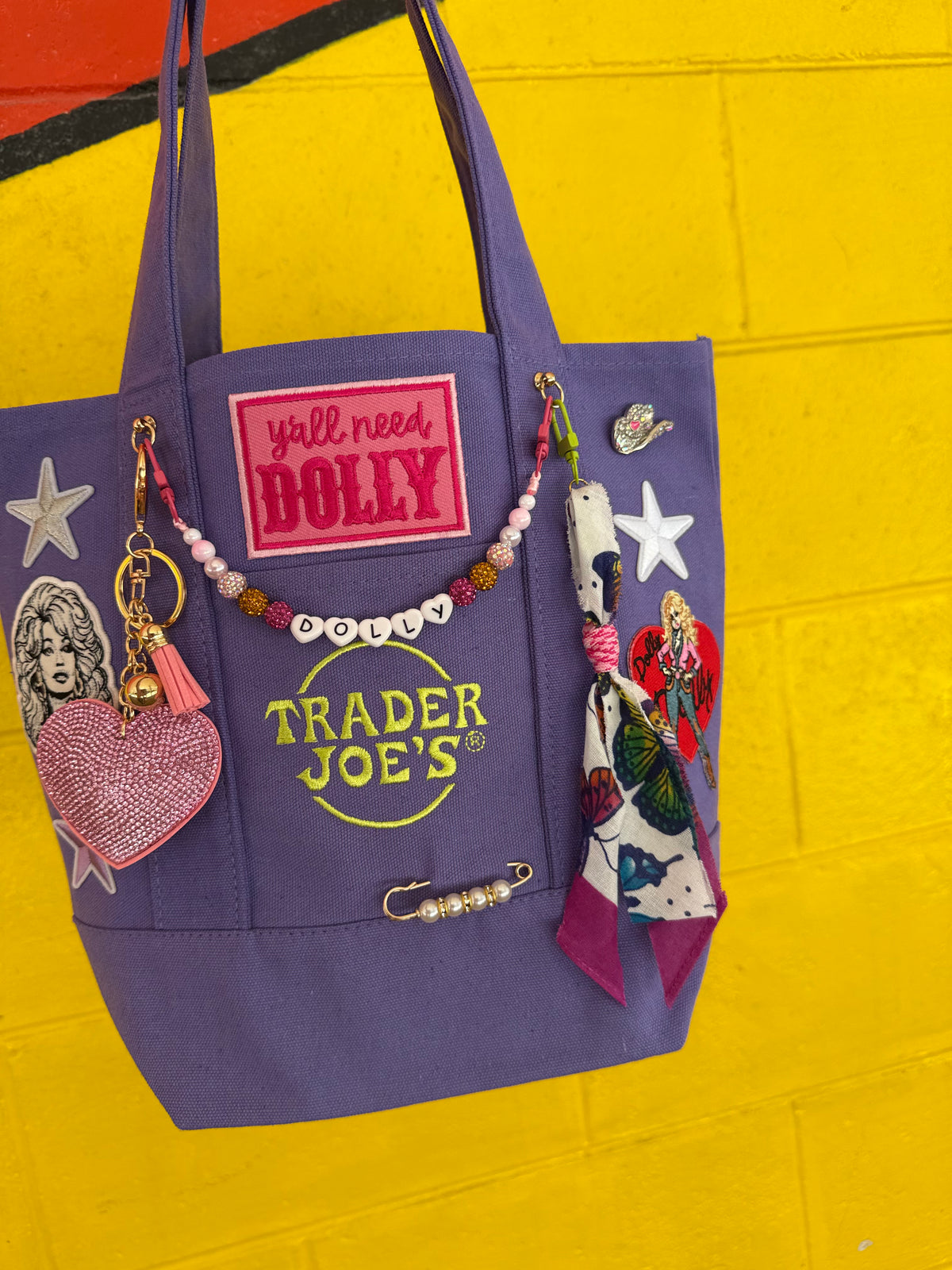 A purple Trader Joe's mini tote decorated with patches, pins, charms and a beaded DOLLY bag chain, hanging against a bright yellow painted brick wall. Dolly Parton Y'all Need Dolly mini tote by PrettyMUCH.
