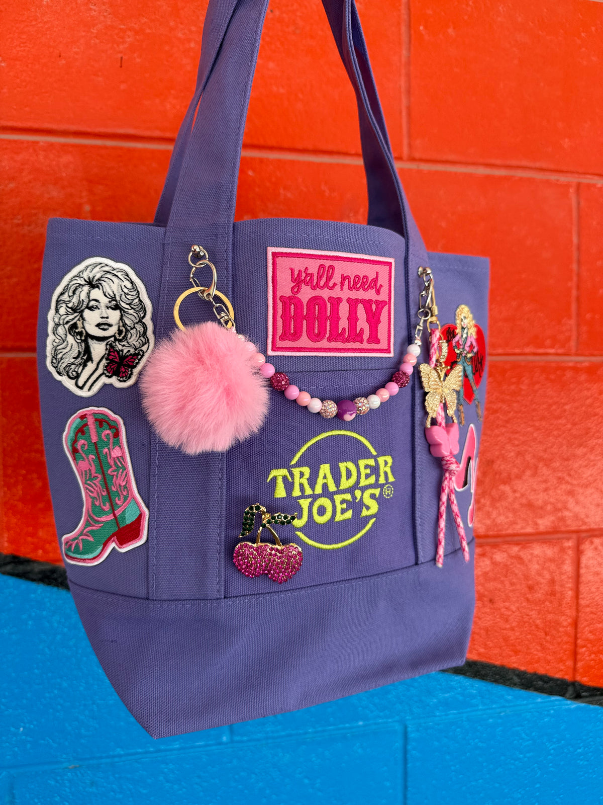 A purple tote bag decorated with various patches, a pink pom-pom, and a beaded keychain, set against a bright orange brick wall and blue floor. Dolly Parton Boots & Heels mini tote by PrettyMUCH.