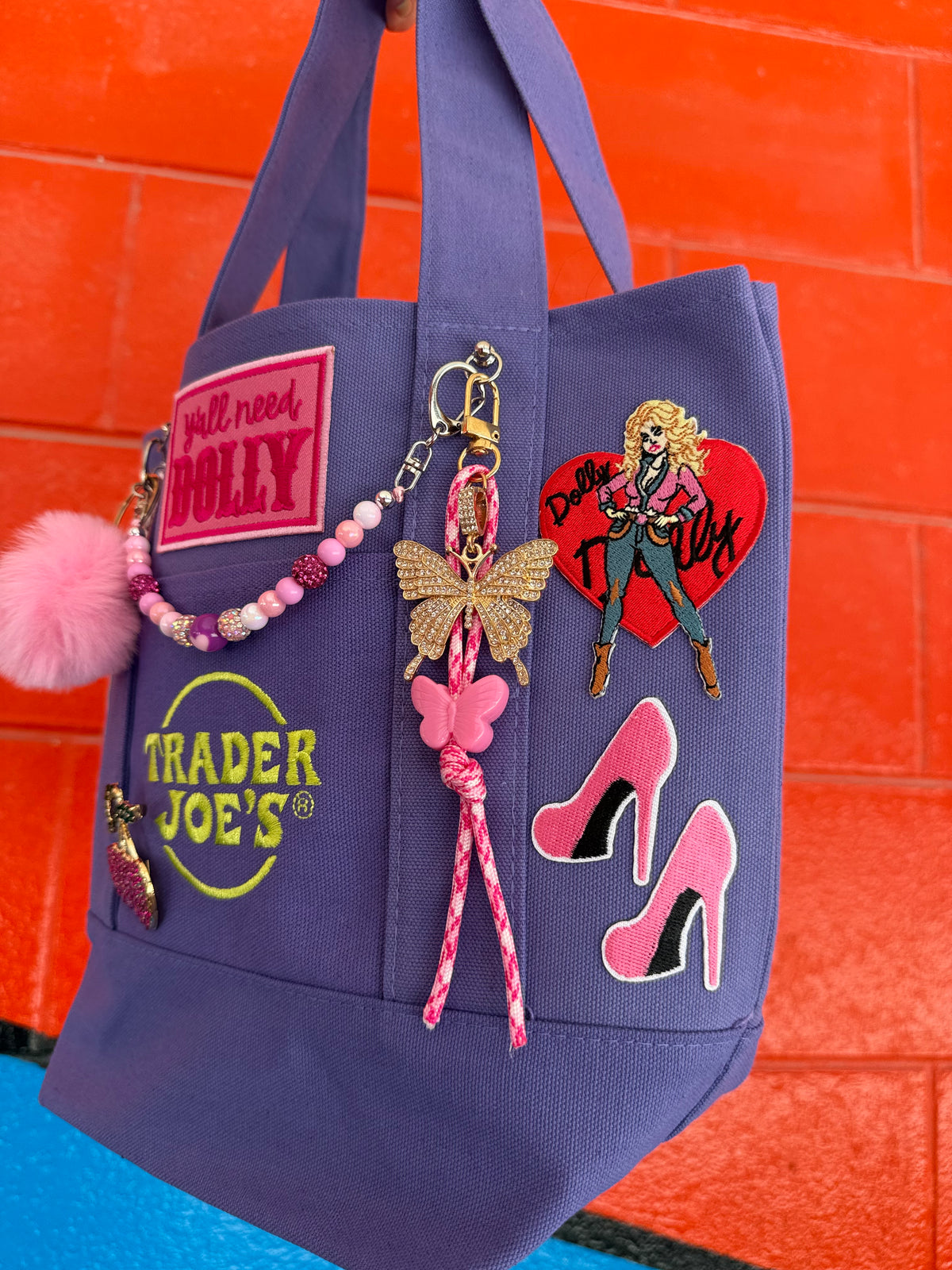 A purple Trader Joe's mini tote with a hot pink 'y'all need DOLLY' patch, the embroidered Trader Joe's logo, pink stiletto patches and a butterfly charm, set against a bright orange brick wall. Dolly Parton Boots & Heels mini tote by PrettyMUCH.