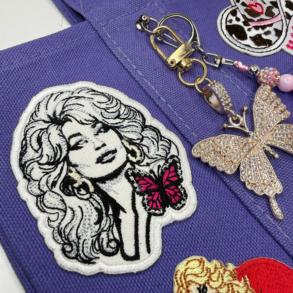 Close-up of the purple Trader Joe's mini tote with the black-and-white embroidered Dolly Parton portrait patch with a pink butterfly, next to a gold rhinestone butterfly keychain. Dolly Parton Sweet but Strong mini tote by PrettyMUCH.