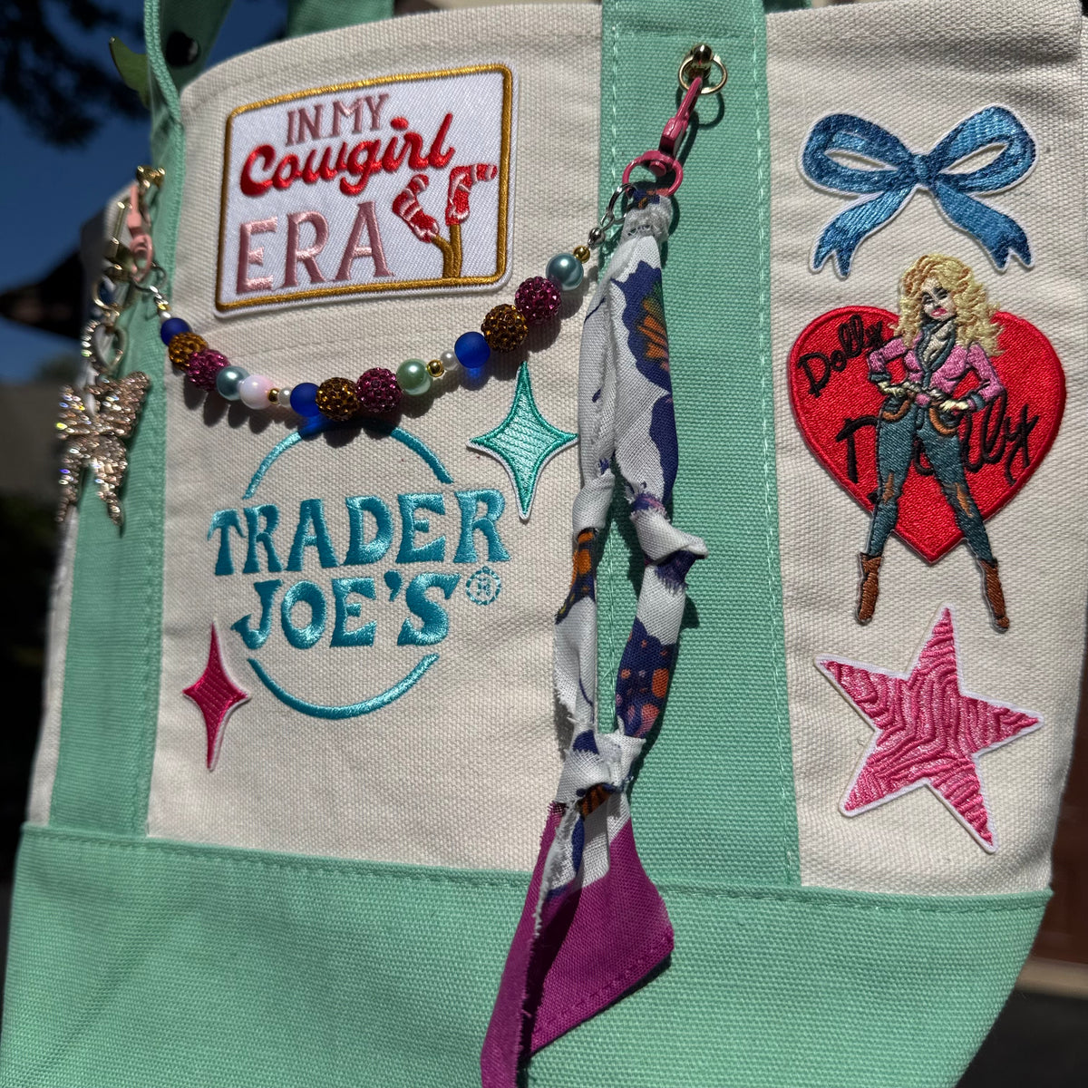 A close-up of a canvas tote bag featuring embroidered patches, including 'IN MY Cowgirl ERA', 'TRADER JOE'S', a cowboy patch, and a heart patch with a cowgirl illustration. A colorful beaded chain and fabric tassels hang from the top. Dolly Parton Cowgirl Era mini tote by PrettyMUCH.