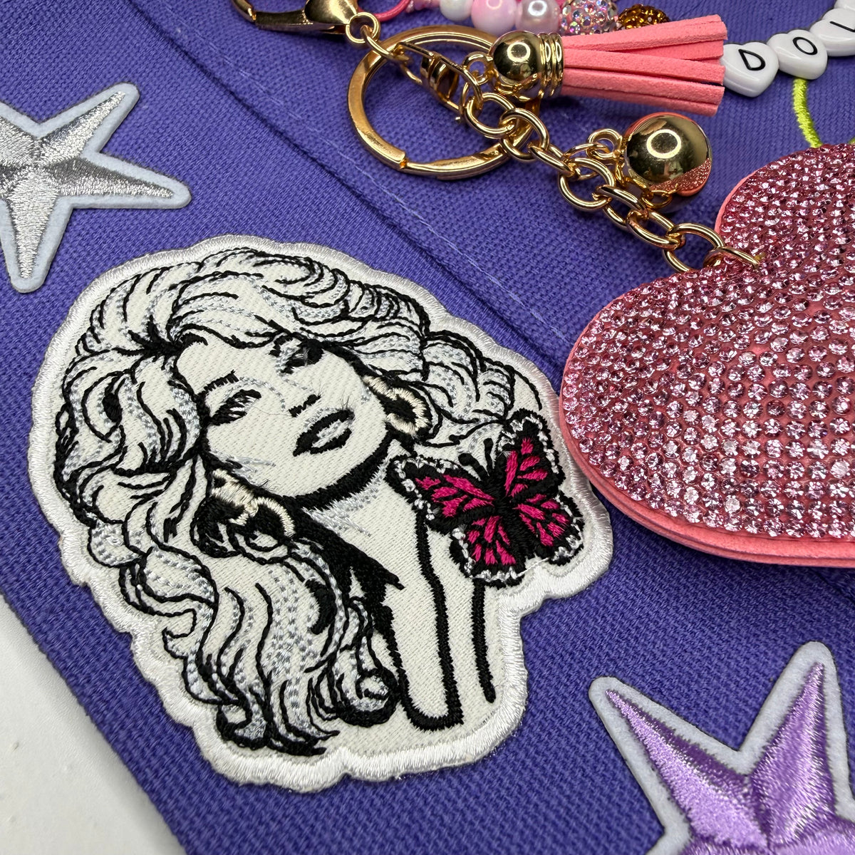 A close-up of a purple fabric item featuring a white embroidered patch with a black-and-white illustrated woman and a pink butterfly, alongside a pink rhinestone heart charm, gold keychain hardware, and decorative star patches. Dolly Parton Y'all Need Dolly mini tote by PrettyMUCH.