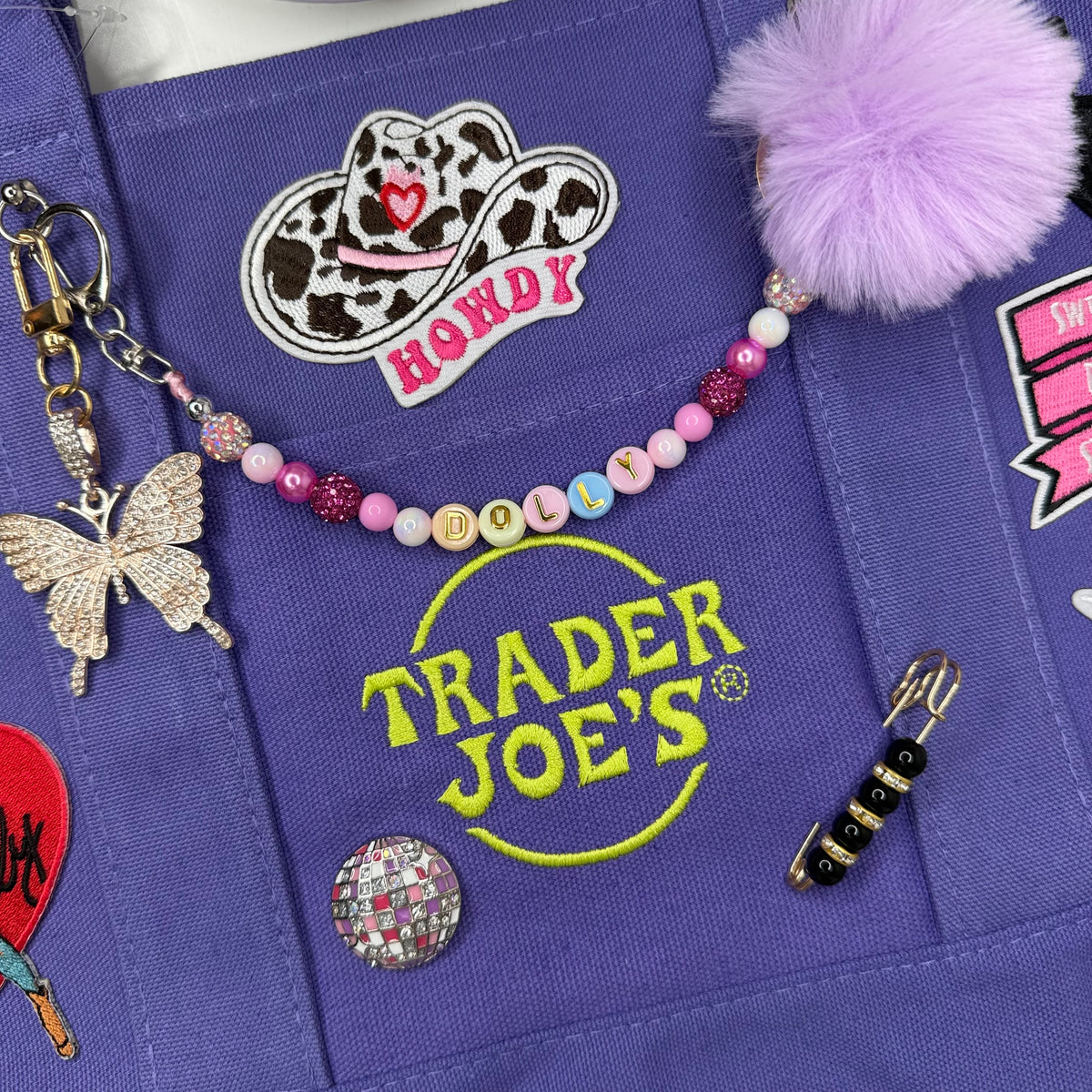 Close-up of the purple Trader Joe's mini tote with the yellow embroidered Trader Joe's logo, a lavender pom-pom, a beaded DOLLY bag chain and a cow-print HOWDY cowgirl hat patch. Dolly Parton Sweet but Strong mini tote by PrettyMUCH.