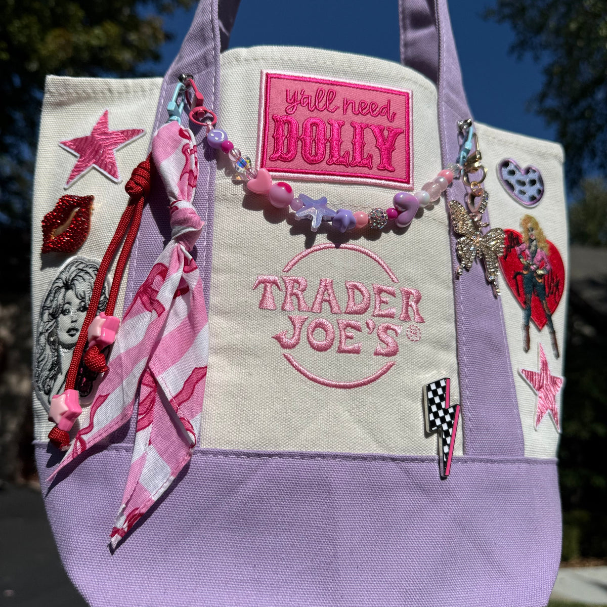 A lavender and cream Trader Joe's mini tote decorated with pink and multicolor patches, a beaded bag chain, a pink-and-white striped bandana twilly and pins, photographed outdoors against a blue sky and green trees. Dolly Parton Pretty in Pastel mini tote by PrettyMUCH.