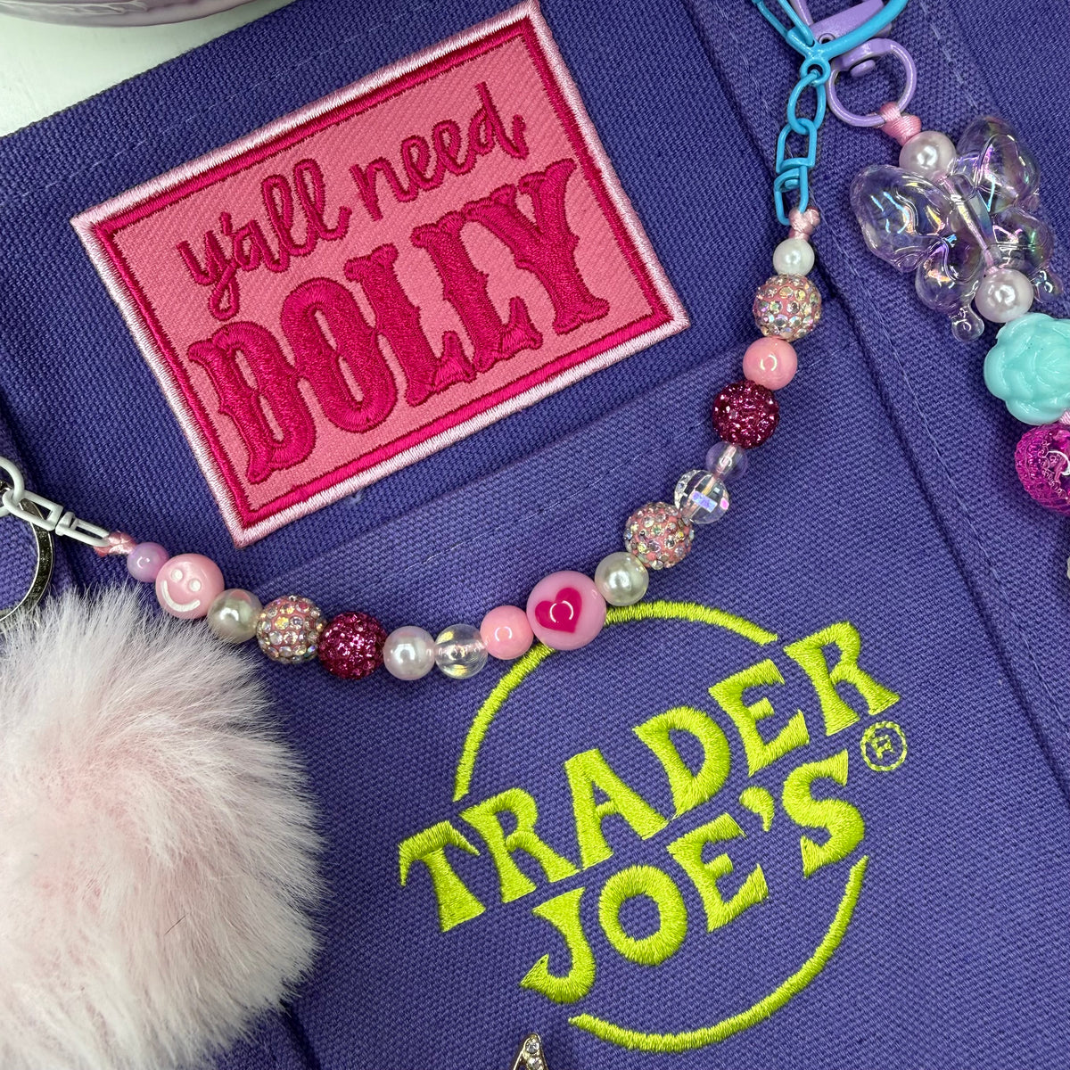 A close-up of a purple fabric accessory featuring a pink embroidered patch that reads 'y'all need DOLLY' and a bright green Trader Joe's logo. A pink beaded charm with pastel beads, a white faux-fur pom-pom, and a butterfly charm are attached. Dolly Parton Aces High mini tote by PrettyMUCH.