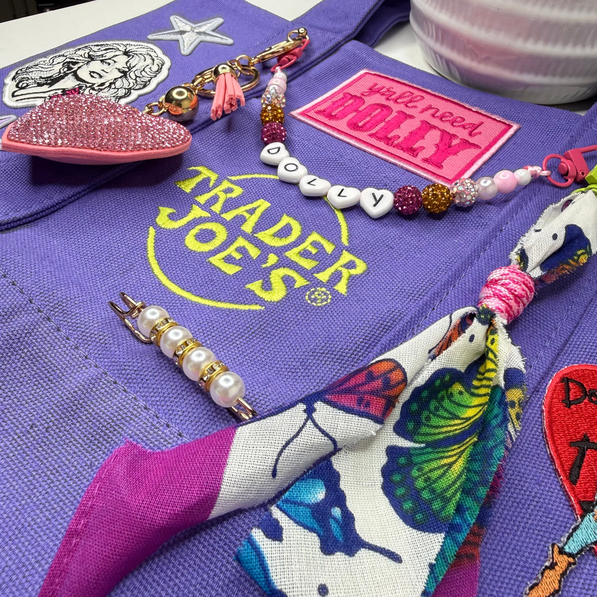 A close-up of a purple fabric accessory, likely a bag or pouch, featuring a yellow Trader Joe's logo patch, a pink 'will need DOLLY' patch, and several colorful charms, beads, and a butterfly-patterned ribbon. Dolly Parton Y'all Need Dolly mini tote by PrettyMUCH.