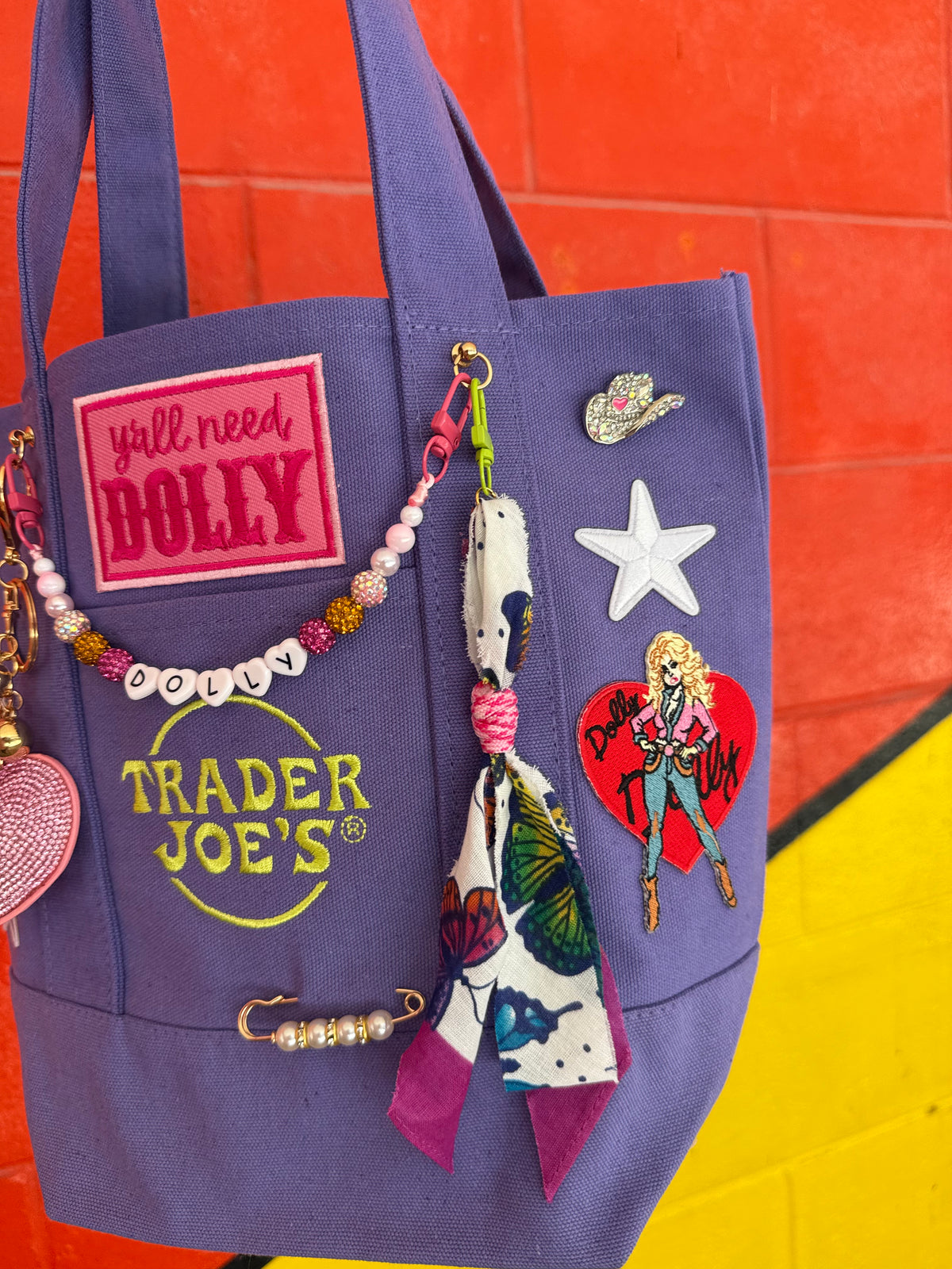 A purple Trader Joe's mini tote with a hot pink 'y'all need DOLLY' patch, the green embroidered Trader Joe's logo, a beaded DOLLY bag chain, a butterfly bandana dangle and a rhinestone cowgirl hat pin, against a bright orange and yellow brick wall. Dolly Parton Y'all Need Dolly mini tote by PrettyMUCH.