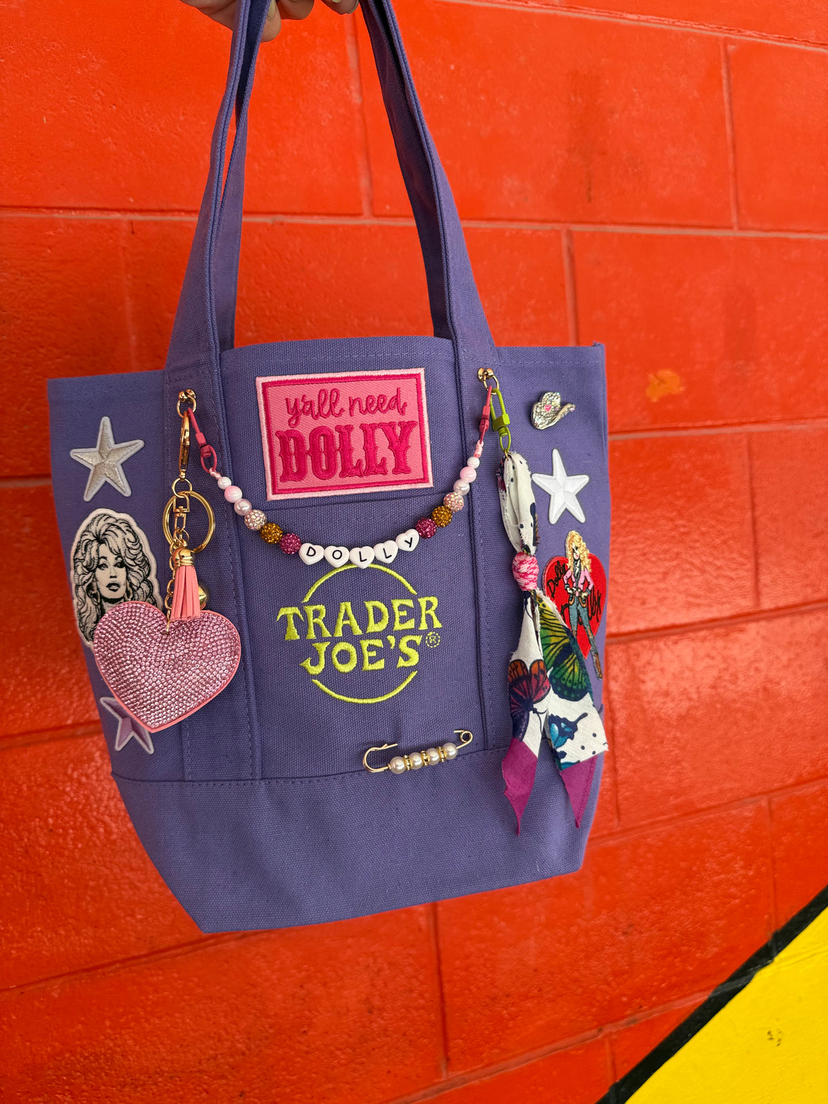A purple Trader Joe's mini tote with embroidered patches, including the hot pink 'y'all need DOLLY' patch and the Trader Joe's logo, hanging against a bright orange brick wall. Dolly Parton Y'all Need Dolly mini tote by PrettyMUCH.