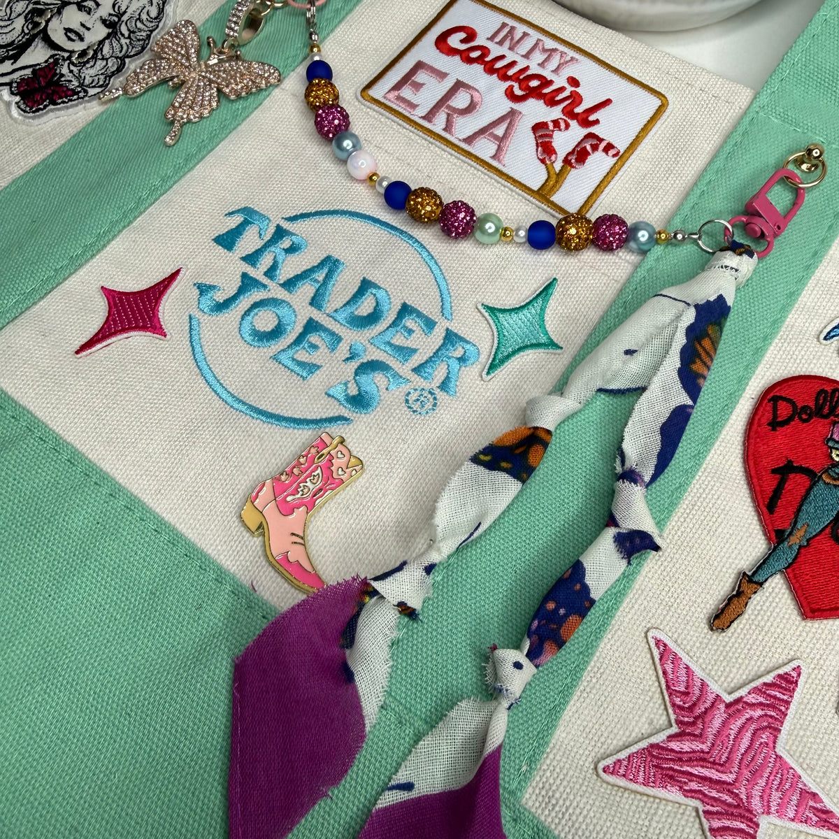 Close-up of the mint and cream Trader Joe's mini tote with the embroidered Trader Joe's logo, an 'In My Cowgirl Era' patch, a gold rhinestone butterfly keychain and a colorful rhinestone bead chain. Dolly Parton Cowgirl Era mini tote by PrettyMUCH.