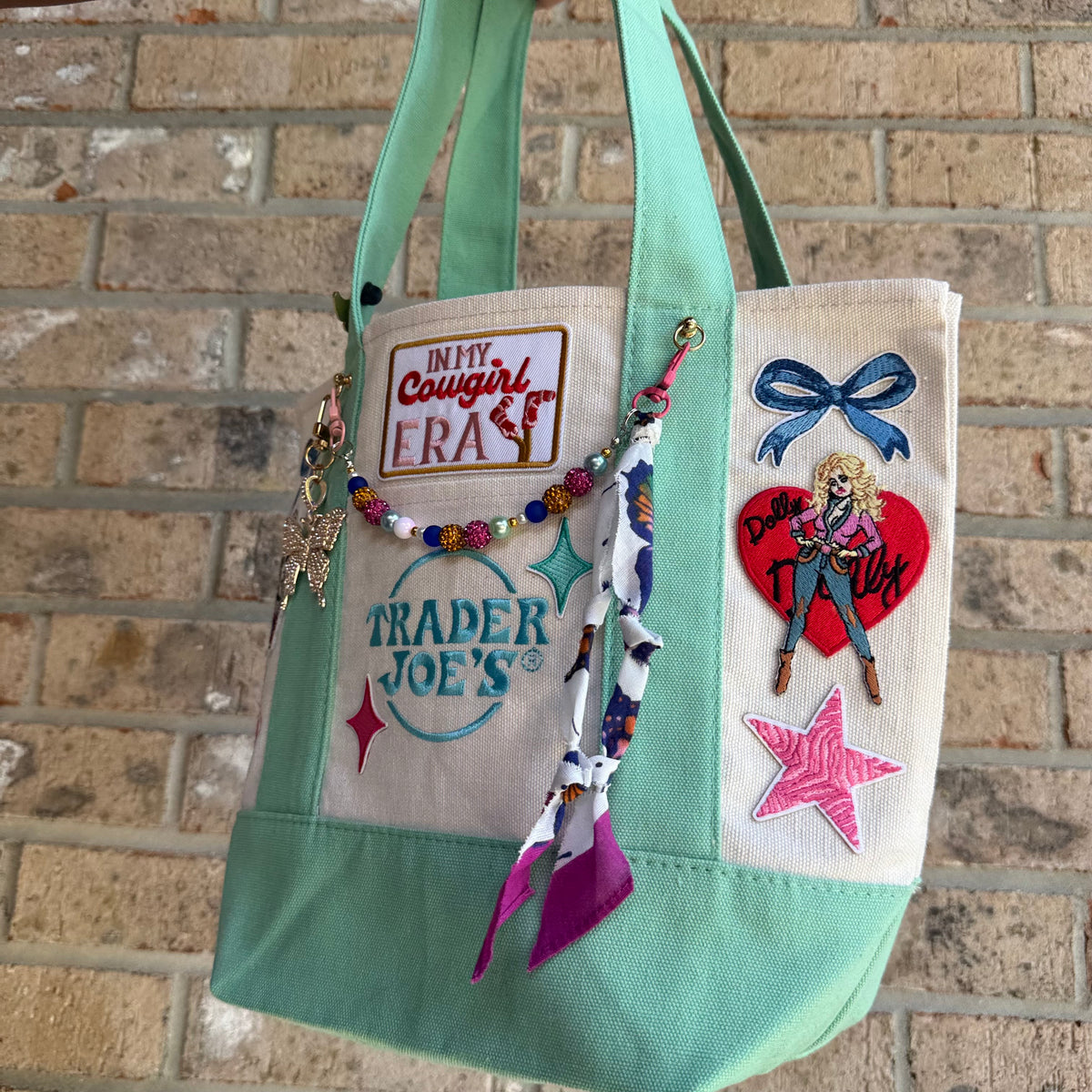 A mint green and cream canvas tote bag decorated with various colorful embroidered patches, a beaded charm strap, and a small metal star charm, hanging against a tan brick wall. Dolly Parton Cowgirl Era mini tote by PrettyMUCH.
