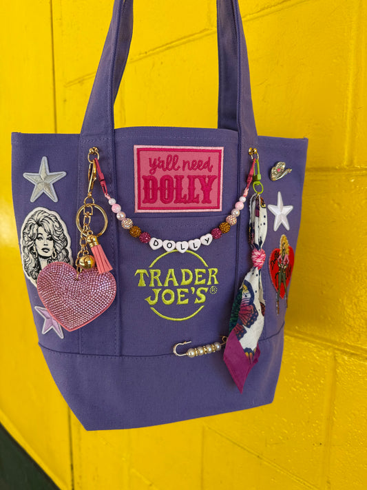 A purple canvas tote bag decorated with various patches, charms, and a beaded necklace, hanging against a bright yellow brick wall. The bag features a pink patch reading 'y'all need DOLLY' and a green 'TRADER JOE'S' logo. Y'all Need Dolly mini custom tote by PrettyMUCH.