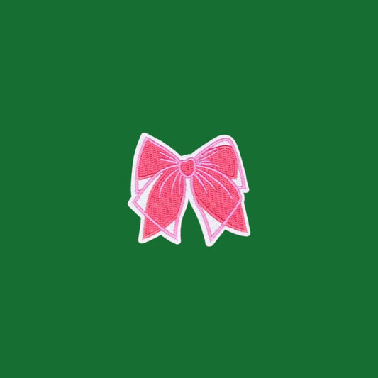 A pink embroidered bow patch with a white border, centered on a solid brown background.