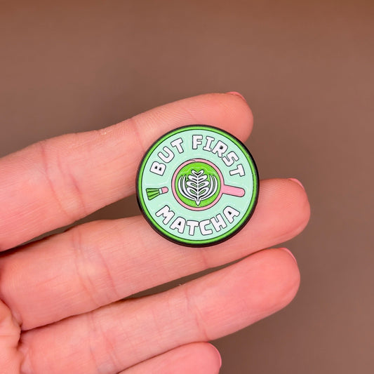 A small round enamel pin held on a person's fingers against a soft brown background. The pin reads 'BUT FIRST MATCHA' with a matcha leaf and green tea droplets design.