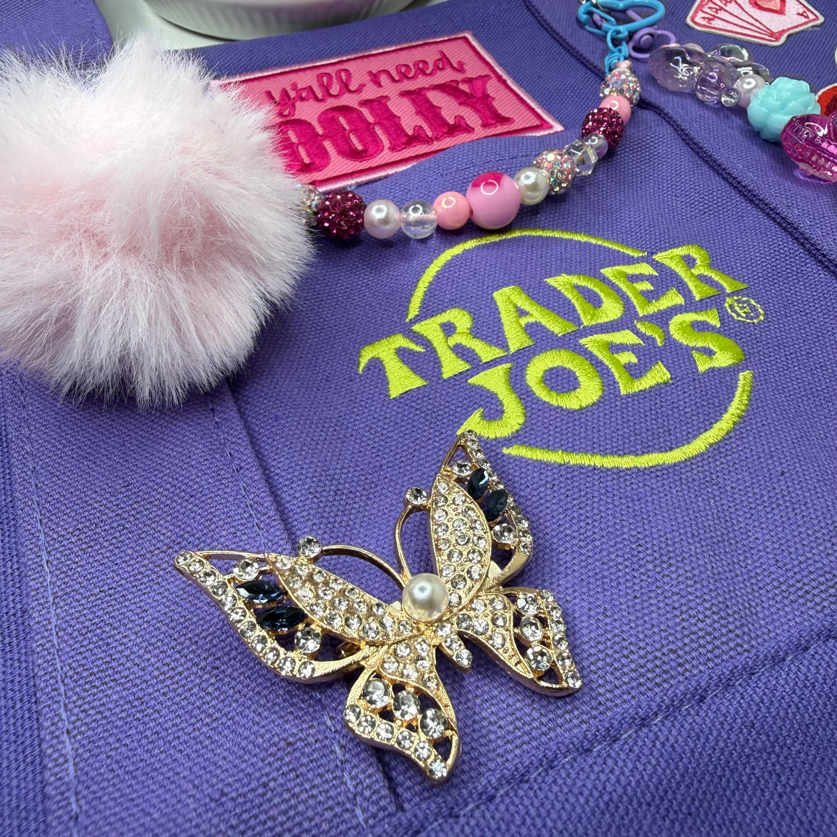Close-up of the purple Trader Joe's mini tote with the embroidered Trader Joe's logo, a pink fuzzy pom-pom, a beaded bag chain and a gold rhinestone butterfly brooch with a pearl center. Dolly Parton Aces High mini tote by PrettyMUCH.
