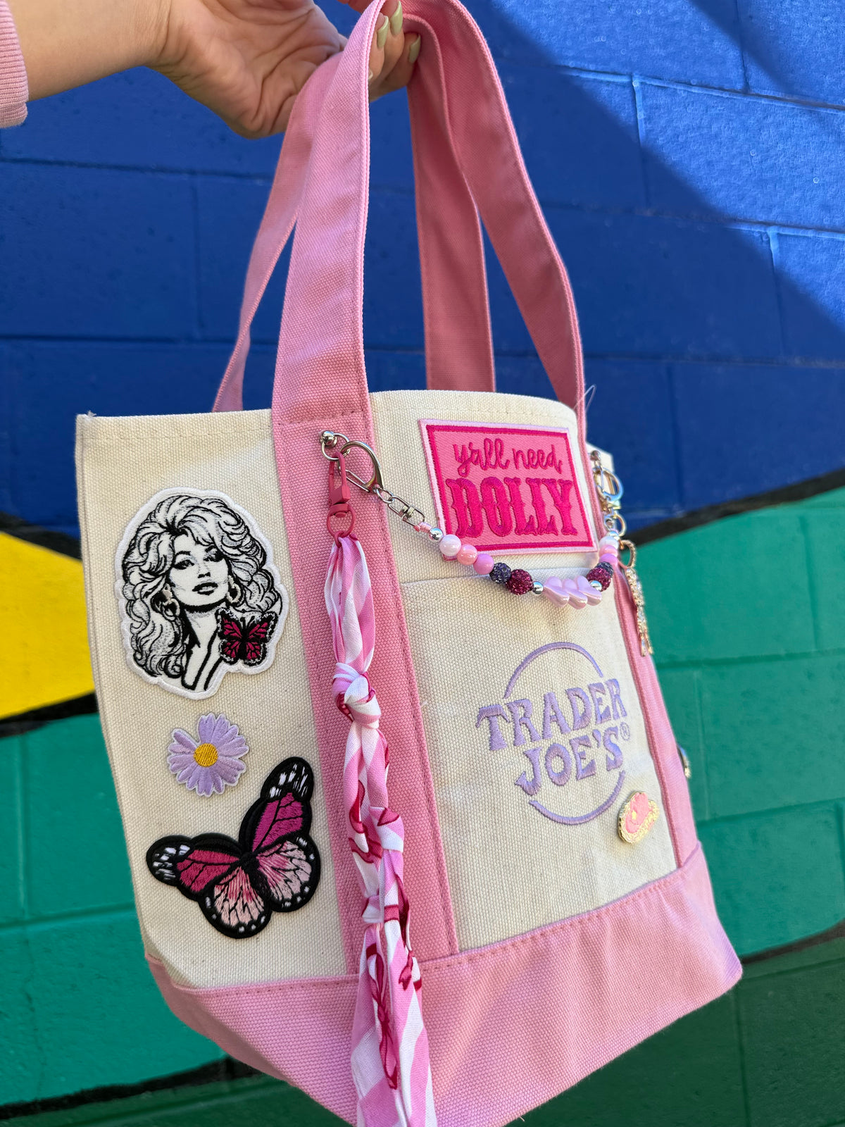 A pink and beige canvas tote bag with embroidered patches, a butterfly applique, and a pink braided tassel charm, held by a hand against a colorful blue and green brick wall. Dolly Parton Pink Bandana mini tote by PrettyMUCH.