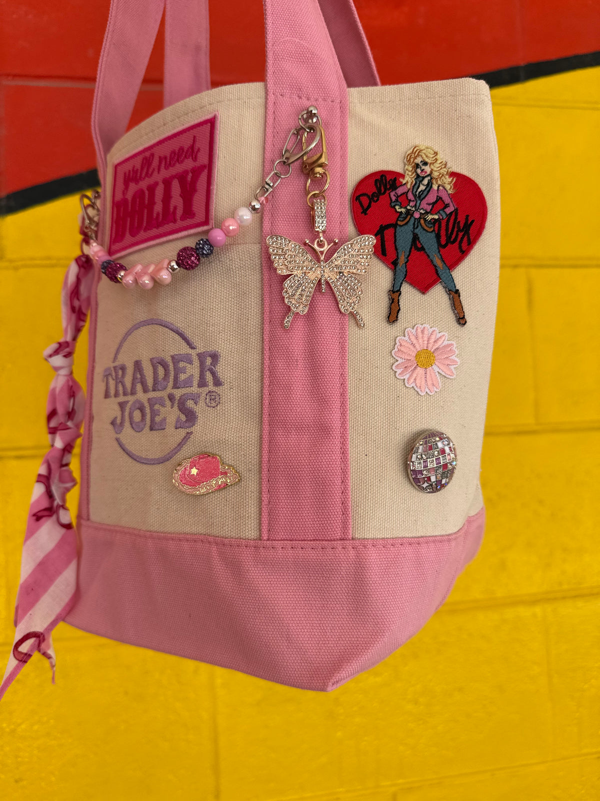 A pink and beige canvas tote bag decorated with various patches, pins, and charms, hanging against a bright yellow and red painted brick wall. Dolly Parton Pink Bandana mini tote by PrettyMUCH.