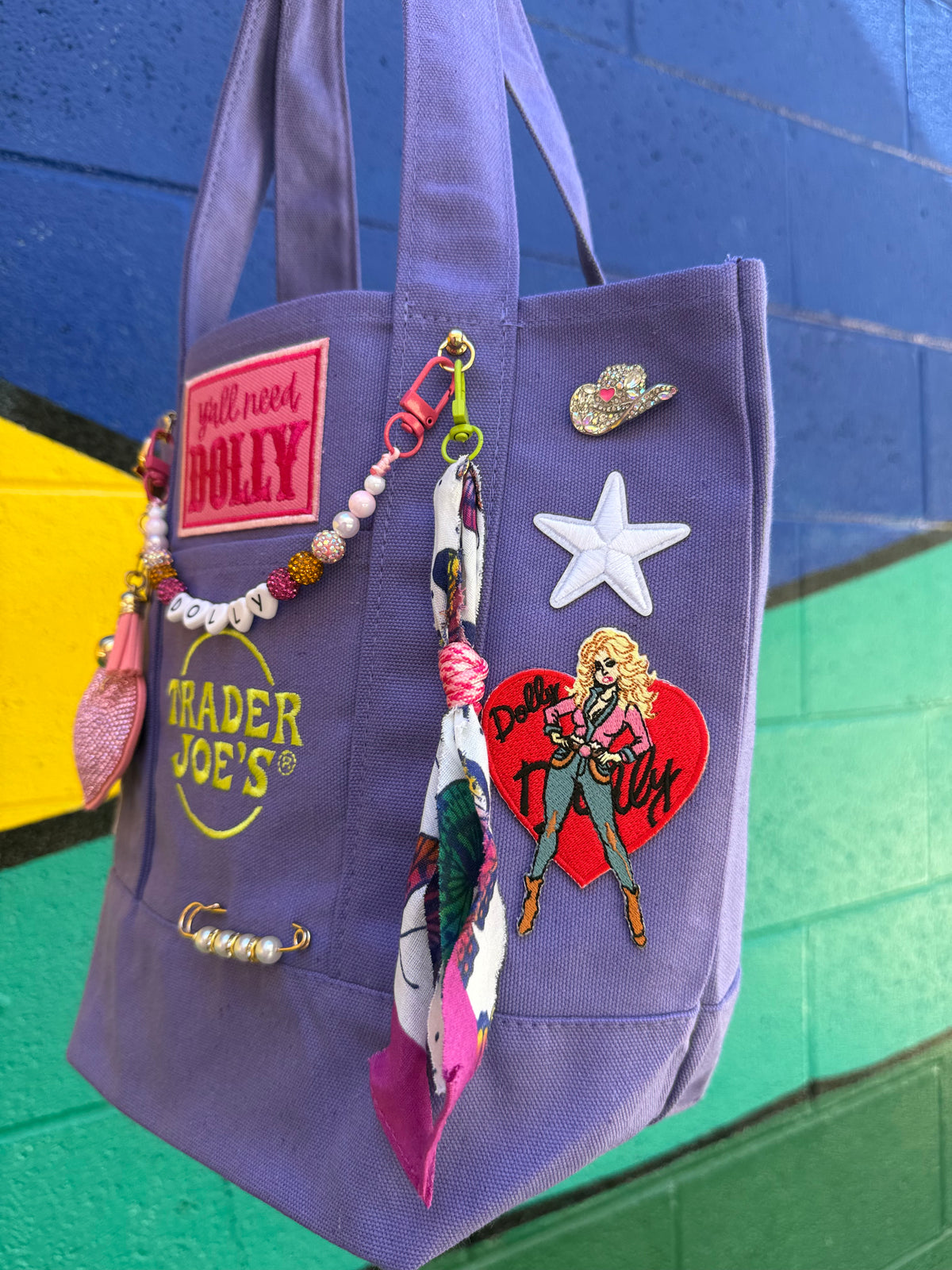 A purple canvas tote bag with various decorative patches, pins, and a beaded keychain strap, hanging against a painted brick wall. Dolly Parton Y'all Need Dolly mini tote by PrettyMUCH.