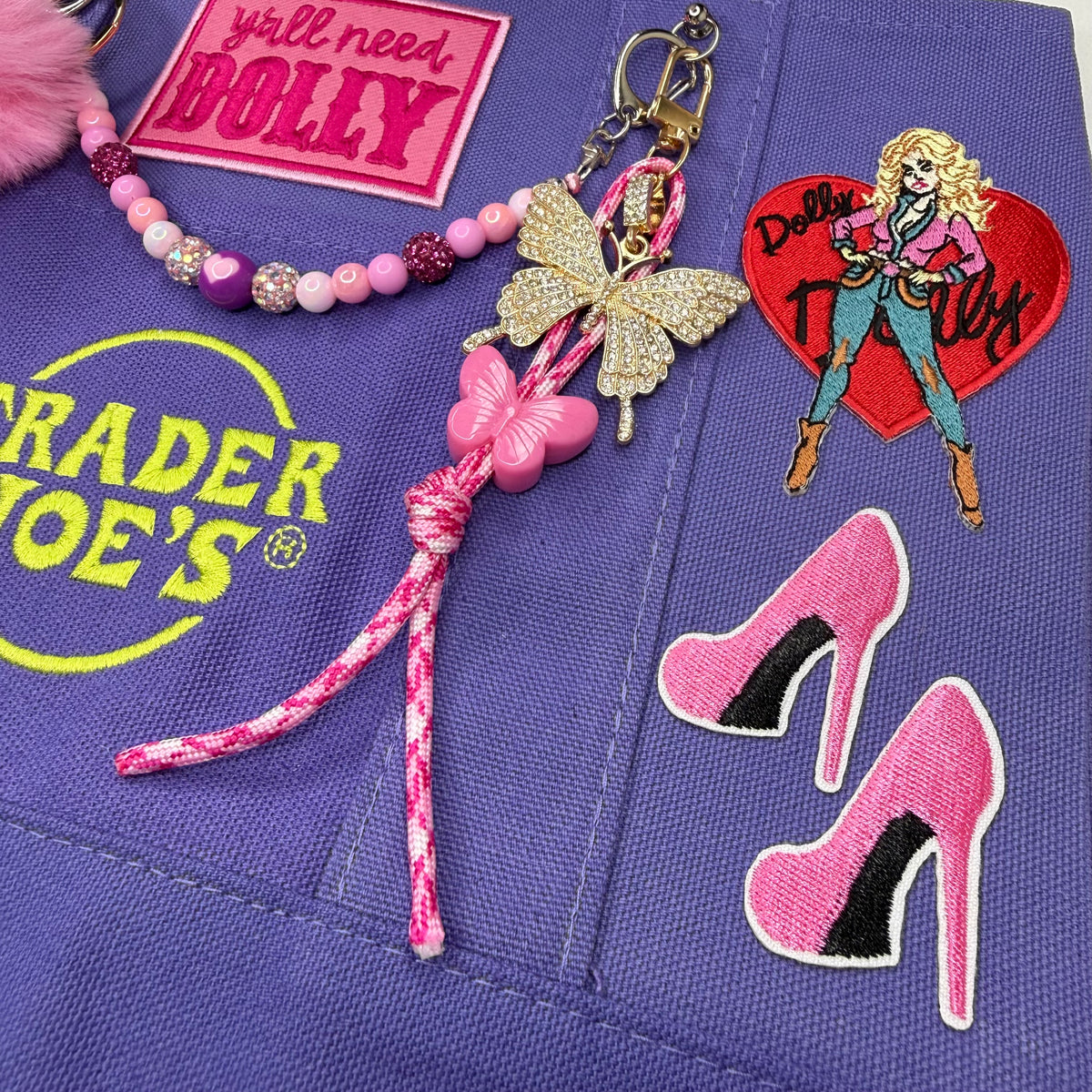 A close-up of a purple fabric accessory featuring embroidered patches and charms inspired by Dolly Parton. The set includes a pink beaded bracelet, a butterfly keychain with a pink bow, a heart patch reading 'Dolly Parton Whoo', a pair of pink high-heel patches, and a yellow Trader Joe's logo patch. Dolly Parton Boots & Heels mini tote by PrettyMUCH.