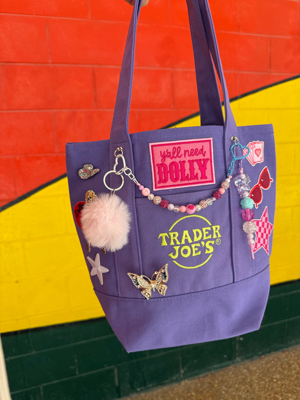 A purple Trader Joe's mini tote decorated with charms, a pink pom-pom and patches, including the embroidered Trader Joe's logo and the hot pink 'y'all need DOLLY' patch, set against a colorful red and yellow brick wall. Dolly Parton Aces High mini tote by PrettyMUCH.