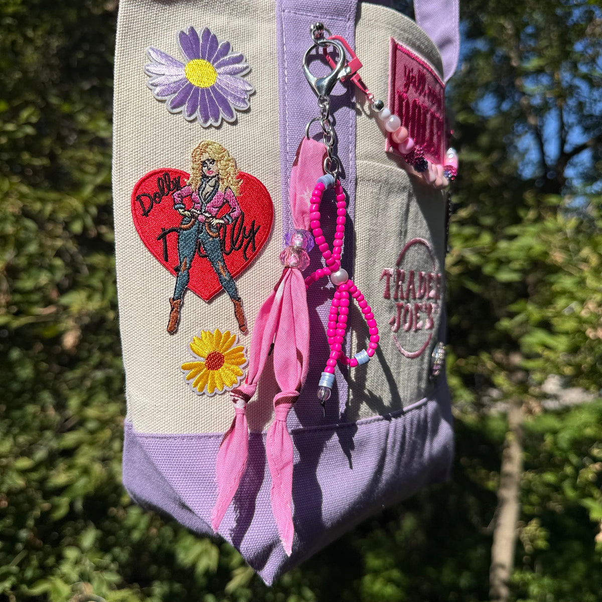 Close-up of the lavender and cream Trader Joe's mini tote decorated with embroidered patches, a pink beaded bag chain and a pink bandana twilly, photographed outdoors against a blurred green background. Dolly Parton You Go Girl mini tote by PrettyMUCH.