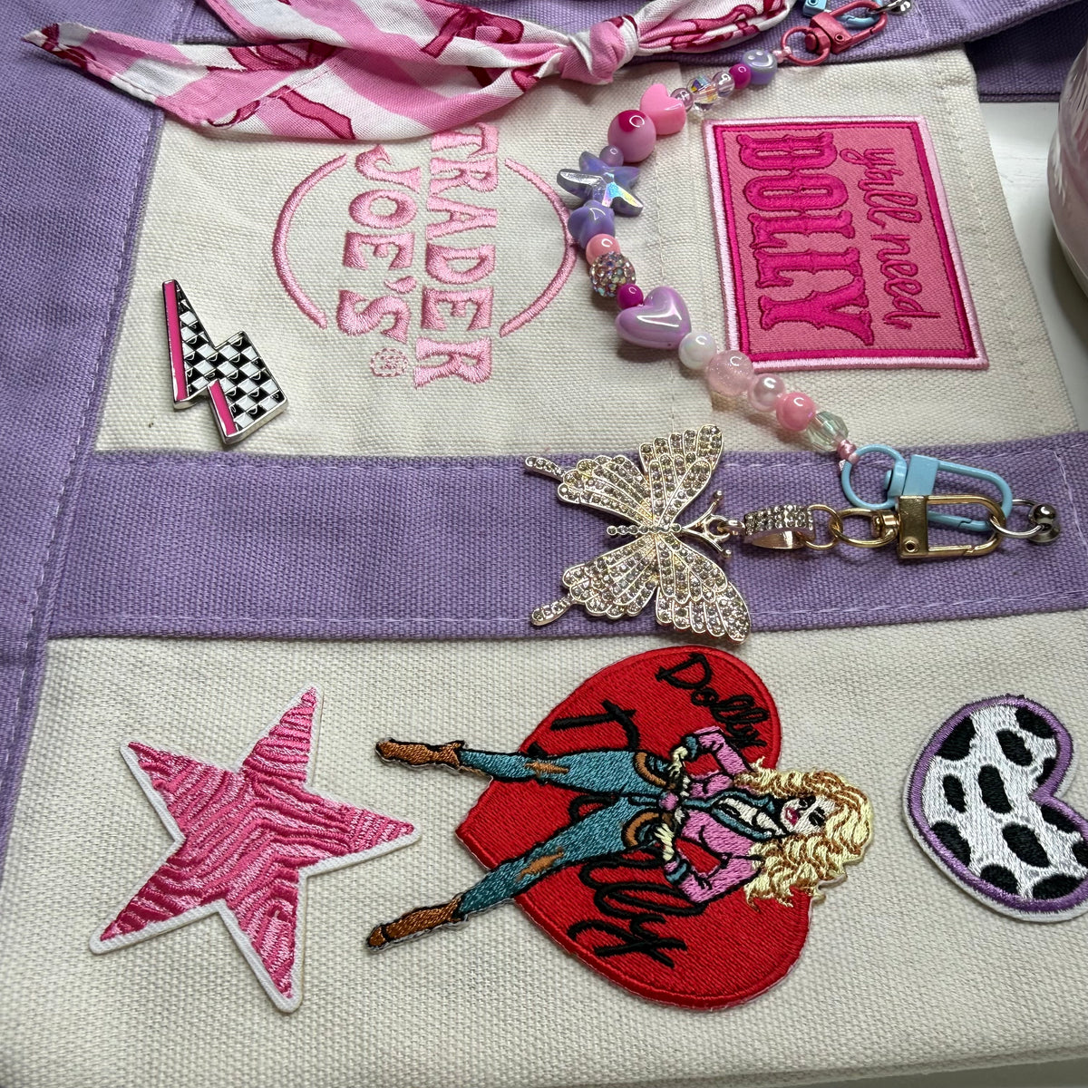 Close-up of the lavender and cream Trader Joe's mini tote with embroidered patches, a pink-and-white striped bandana twilly and a beaded bag chain with a butterfly keychain. Dolly Parton Pretty in Pastel mini tote by PrettyMUCH.
