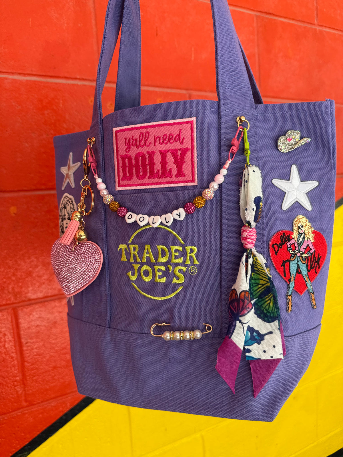 A purple canvas tote bag decorated with various colorful patches, pins, charms, and a small scarf, hanging against a bright orange brick wall with a yellow floor visible at the bottom. Dolly Parton Y'all Need Dolly mini tote by PrettyMUCH.