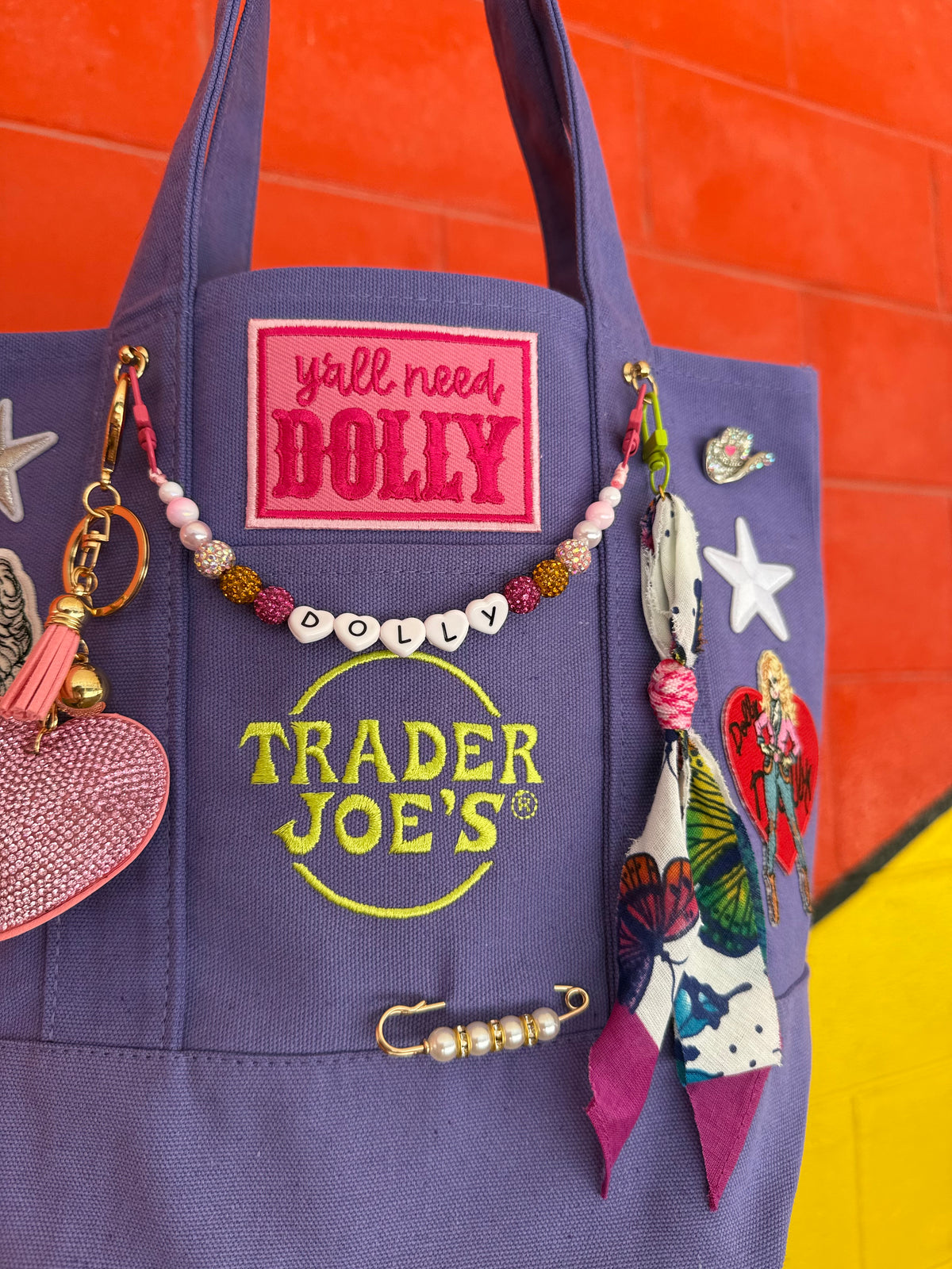 A purple canvas tote bag with embroidered 'Trader Joe's' branding and a pink 'y'all need DOLLY' patch, decorated with colorful beaded jewelry, a pink heart keychain, and fabric charms, set against a bright orange and yellow background. Dolly Parton Y'all Need Dolly mini tote by PrettyMUCH.