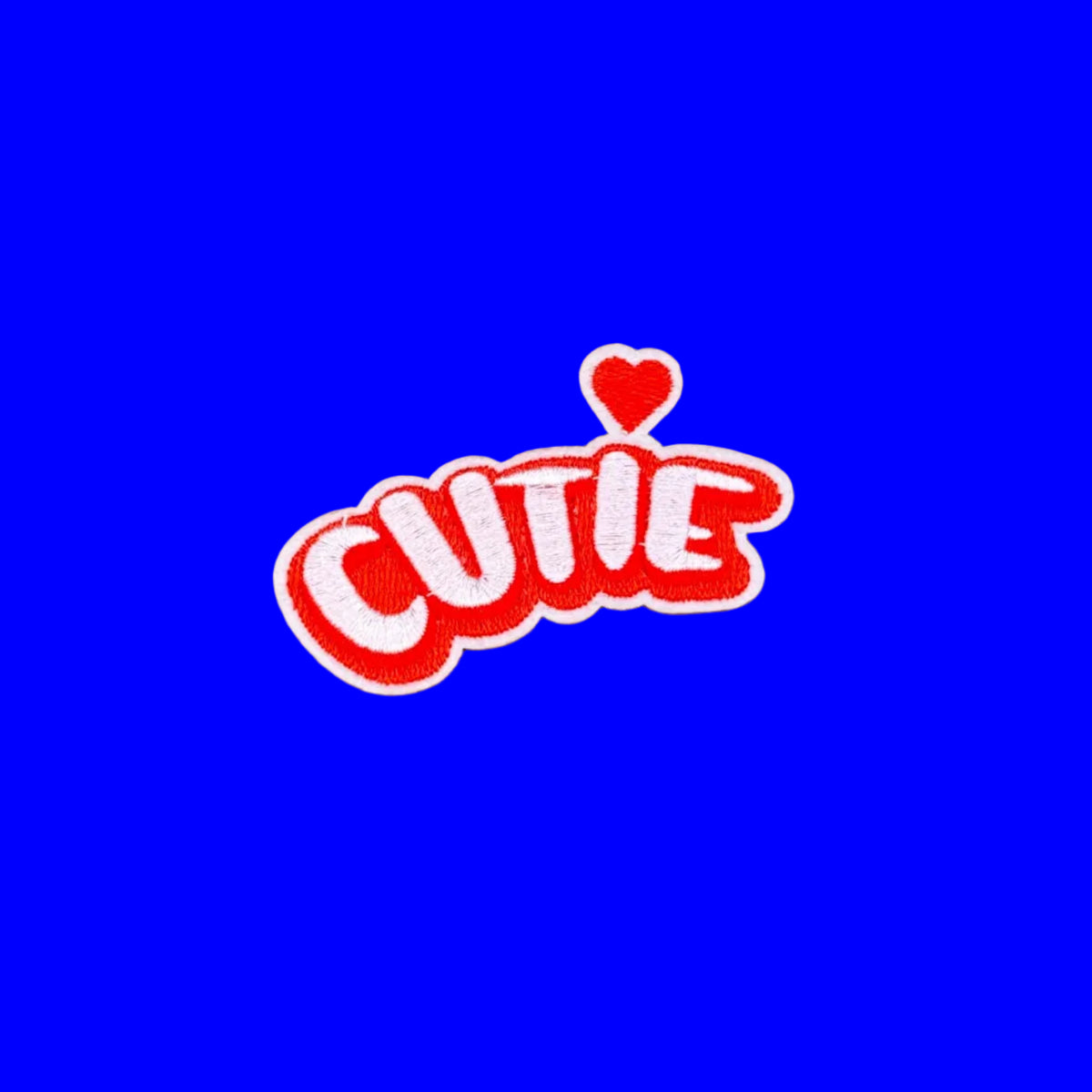 A red and white embroidered patch spelling 'CUTIE' with a small red heart above the letters, placed on a brown textured background.