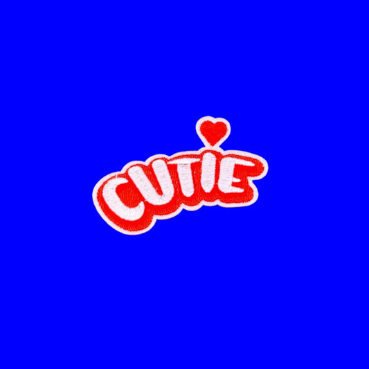 A red and white embroidered patch spelling 'CUTIE' with a small red heart above the letters, placed on a brown textured background.