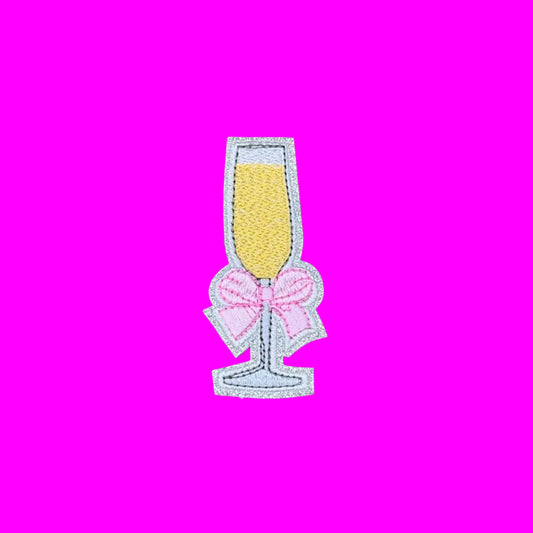An embroidered patch shaped like a champagne flute filled with yellow champagne and decorated with a pink bow, shown on a solid brown background.