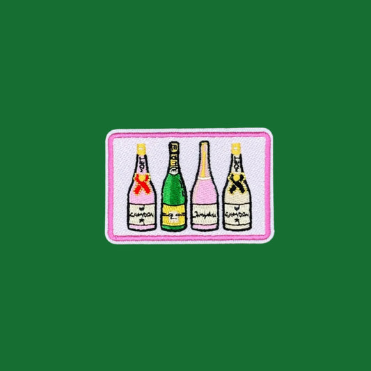 A rectangular embroidered patch featuring four champagne bottles in different colors (green, pink, yellow, black) on a white background with a pink border, displayed on a solid green surface.
