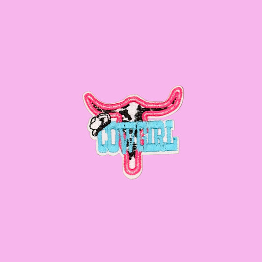 A small embroidered patch featuring a pink longhorn steer silhouette with a cow head detail and the word "COVER" in light blue letters, shown on a plain brown background.