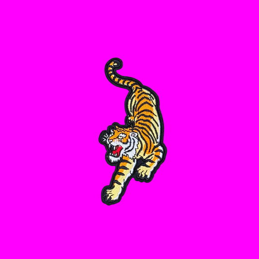 An embroidered iron-on patch of a roaring tiger in yellow and black stripes with a white face, shown on a dusty pink background.