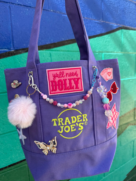 A purple Trader Joe's mini tote decorated with patches, charms and a beaded bag chain, including the hot pink 'y'all need DOLLY' patch, set against a painted green and blue brick wall. Dolly Parton Aces High mini tote by PrettyMUCH.
