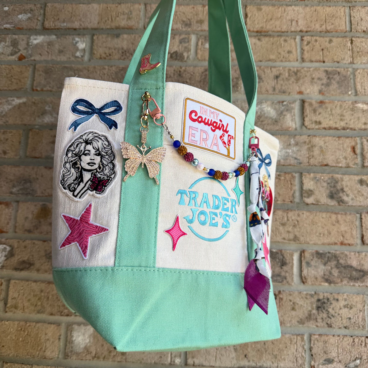 A canvas tote bag with mint green handles and bottom panel, decorated with multiple embroidered patches, a beaded charm, and butterfly embellishments, hanging against a brick wall. Dolly Parton Cowgirl Era mini tote by PrettyMUCH.