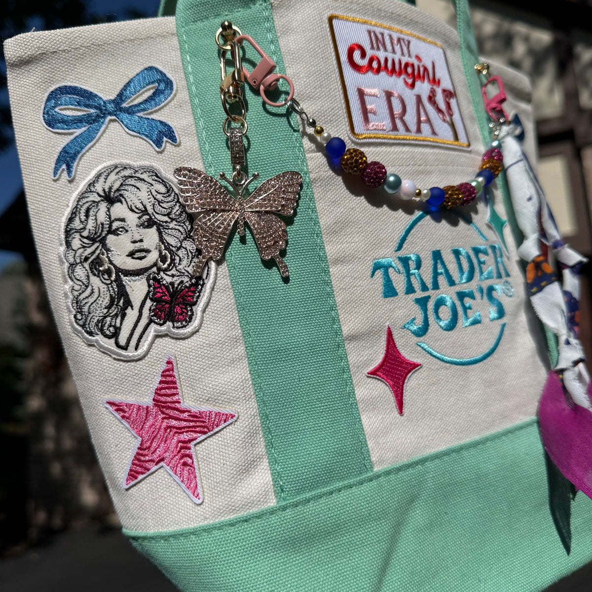A close-up of a canvas tote bag featuring mint green and off-white panels, decorated with colorful embroidered patches, a butterfly charm, and a beaded keychain. Visible text includes 'In My Cowgirl Era' and 'Trader Joe's'. Dolly Parton Cowgirl Era mini tote by PrettyMUCH.