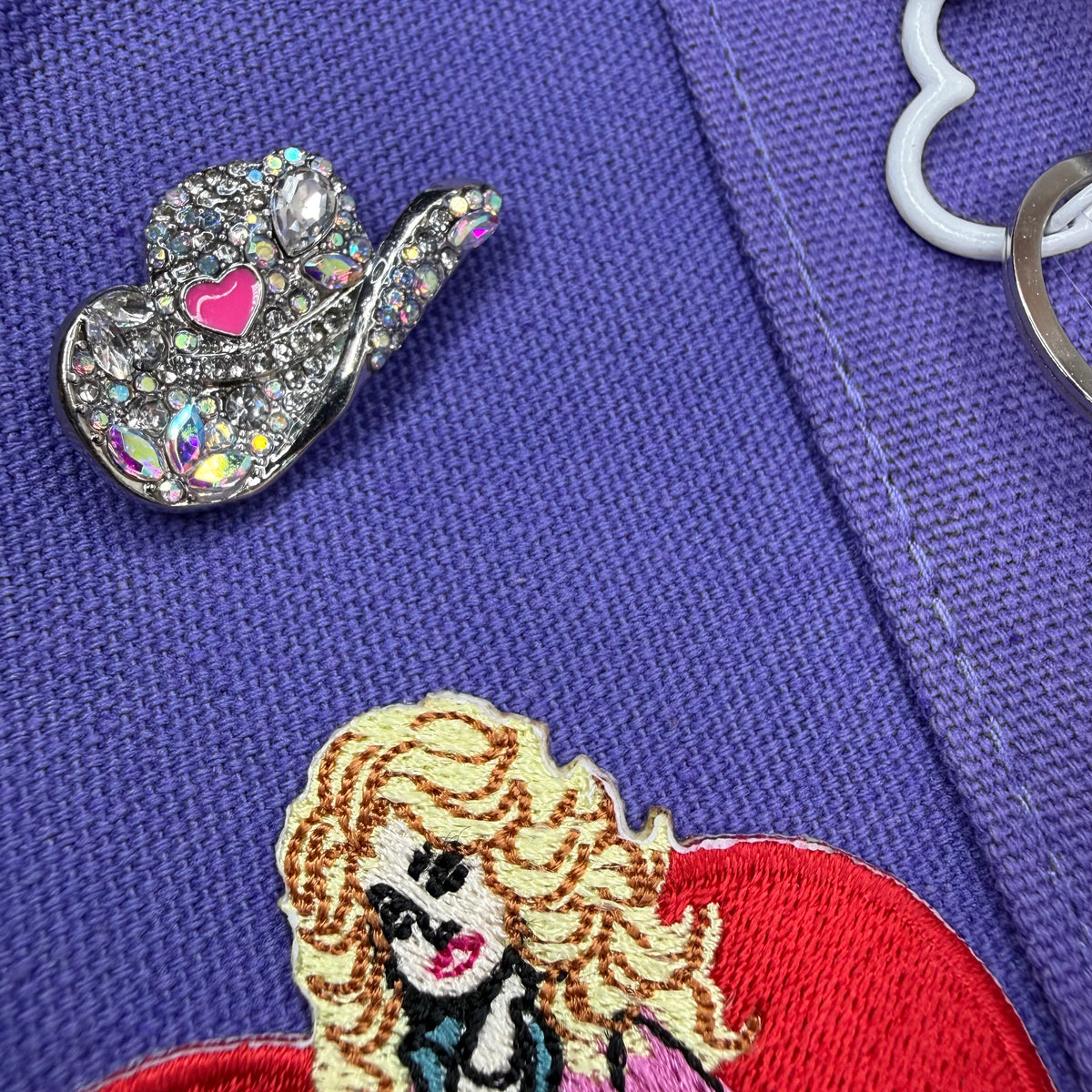 Close-up of the purple Trader Joe's mini tote with a silver rhinestone cowgirl hat pin with a pink heart next to the embroidered Dolly Parton red heart patch. Dolly Parton Aces High mini tote by PrettyMUCH.