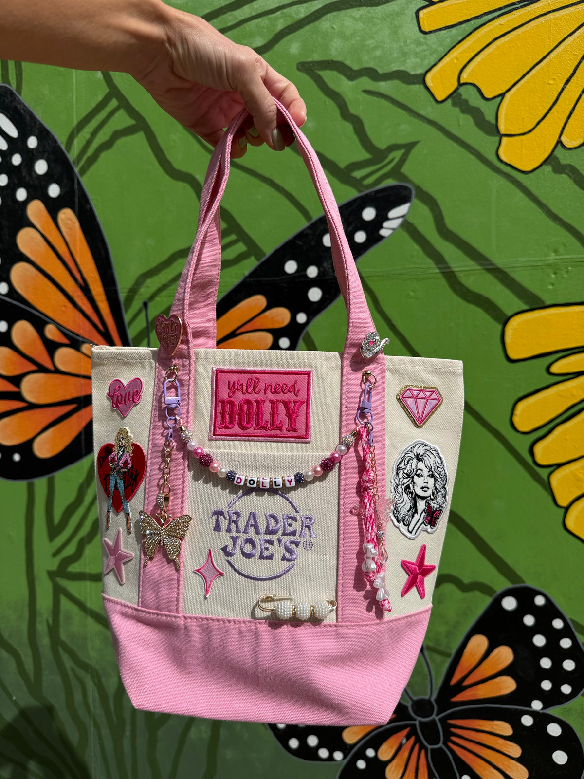 Pink tote decorated with Y'all Need Dolly and Dolly Parton iron-on patches in front of a butterfly mural