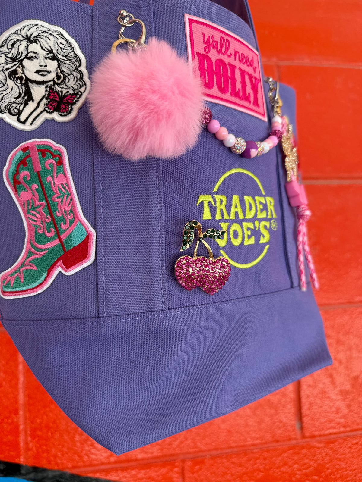 A purple Trader Joe's mini tote decorated with Dolly patches, a pink pom-pom, a beaded bag chain and a rhinestone cherry heart brooch, set against a bright orange brick wall. Dolly Parton Boots & Heels mini tote by PrettyMUCH.