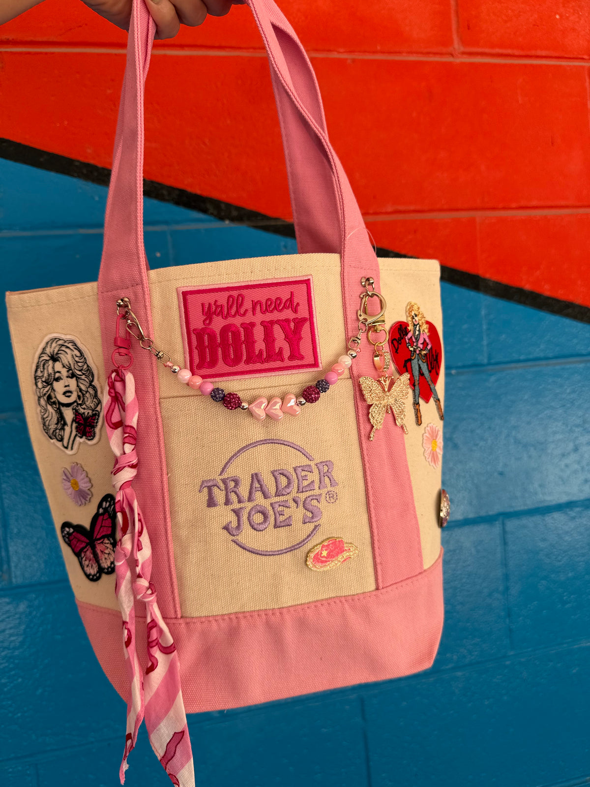 A pink and beige canvas tote bag with multiple decorative patches, embroidered text, and charms, held by a hand against a colorful blue and orange brick wall. Dolly Parton Pink Bandana mini tote by PrettyMUCH.