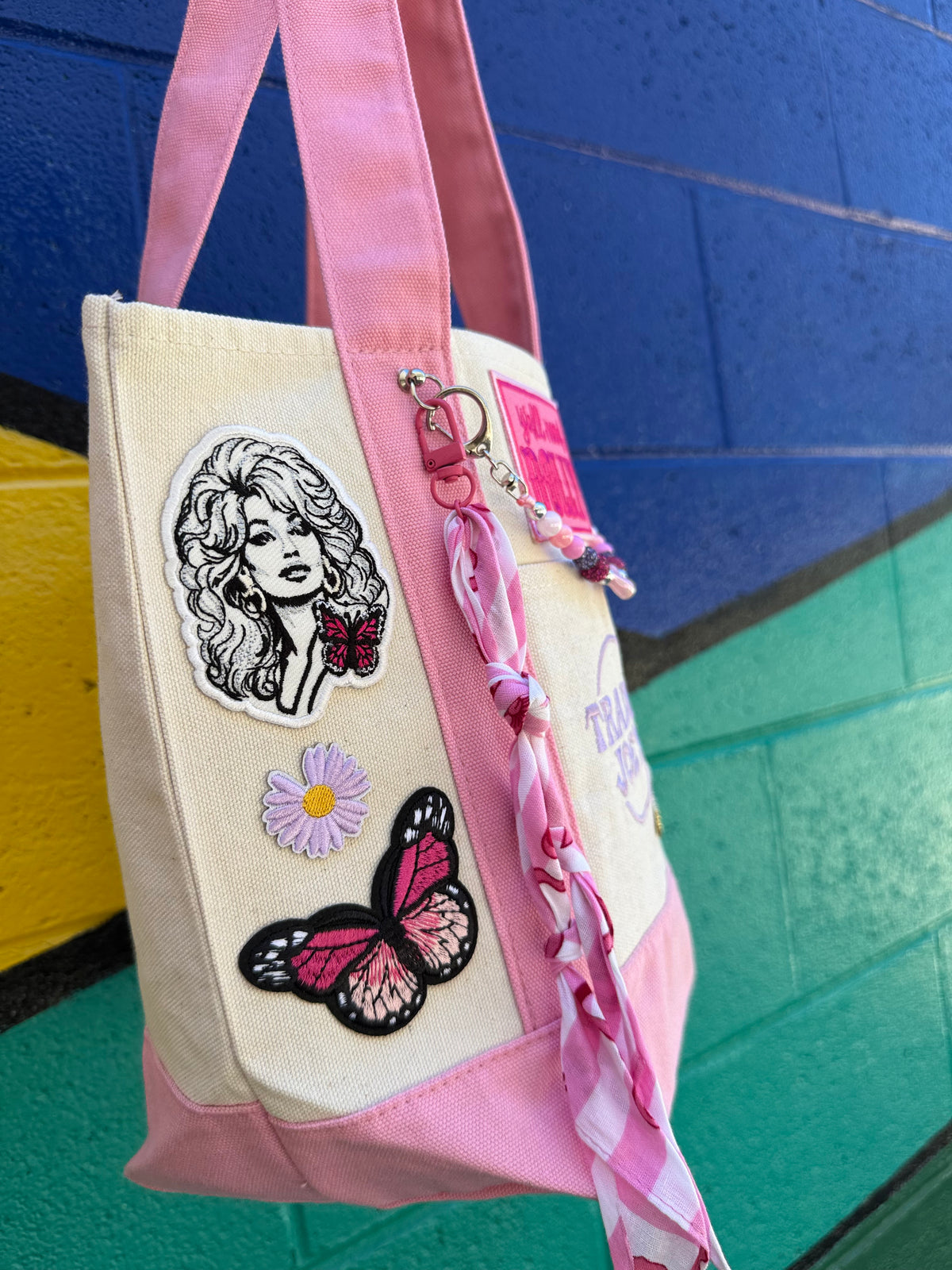 A pink and cream canvas tote bag decorated with embroidered patches, including a butterfly, a daisy, and a portrait patch, hanging against a colorful painted brick wall. Dolly Parton Pink Bandana mini tote by PrettyMUCH.
