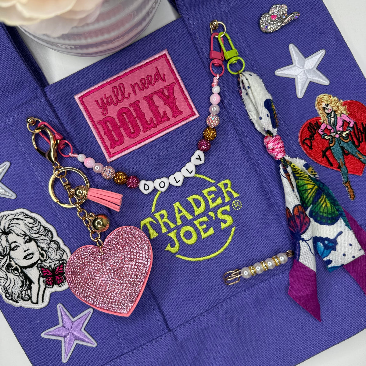 A purple fabric tote bag with embroidered patches and stickers, styled with a beaded keychain, tassel charm, and accessories. Dolly Parton Y'all Need Dolly mini tote by PrettyMUCH.